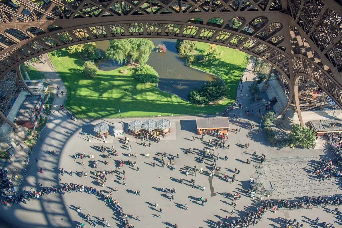 Visitors on the esplanade beneath the Eiffel Tower seen from above, with queue lines along a pillar, daytime