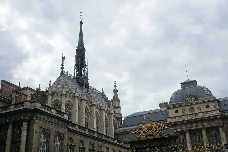 The spire and slate roof of the Sainte-Chapelle rising beside the gilded gate of the Palais de Justice under a cloudy sky