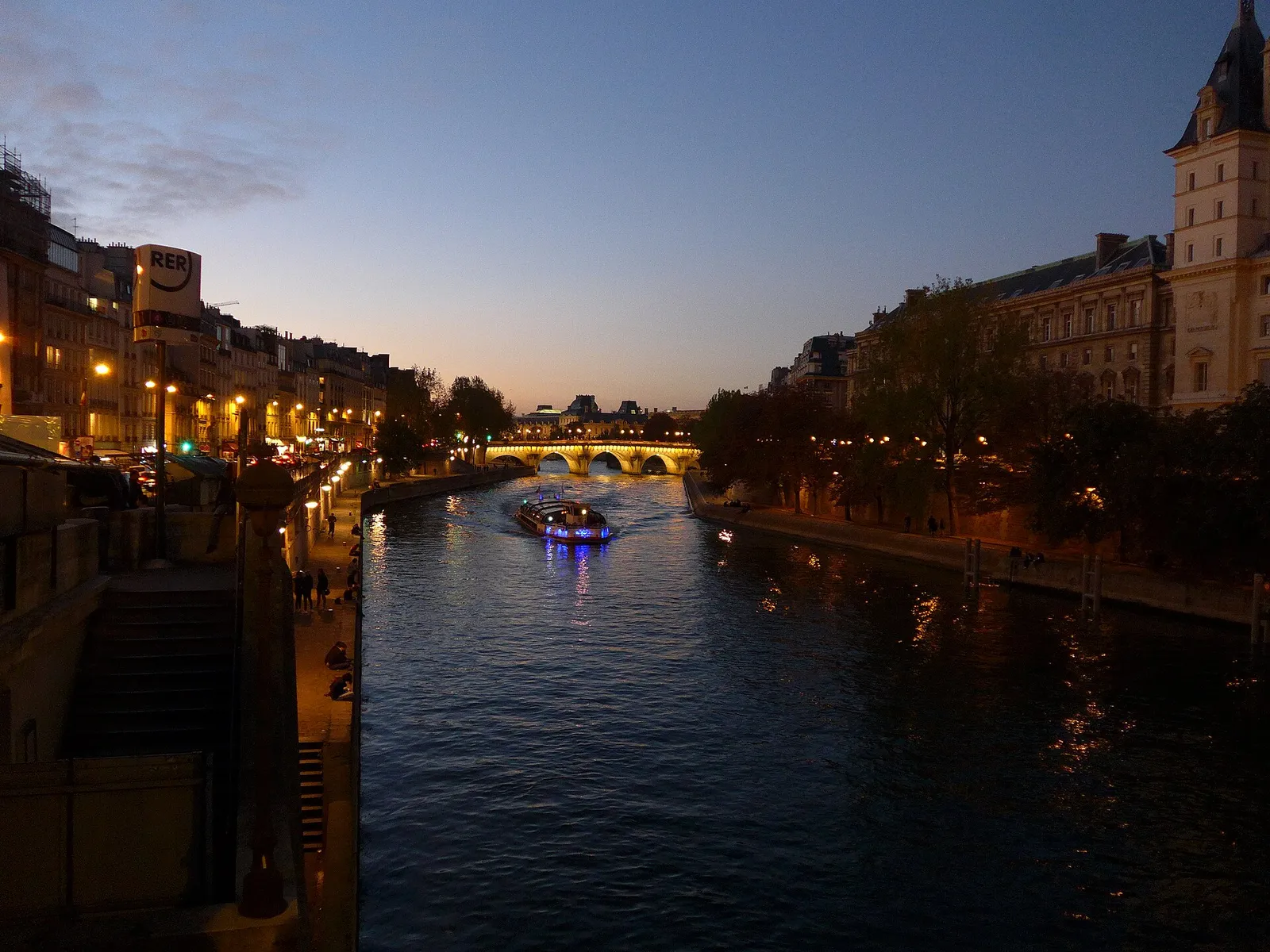 Sightseeing boat with blue lights heading toward the lit Pont Neuf, Seine between the Quai des Orfèvres and the Left Bank, dusk