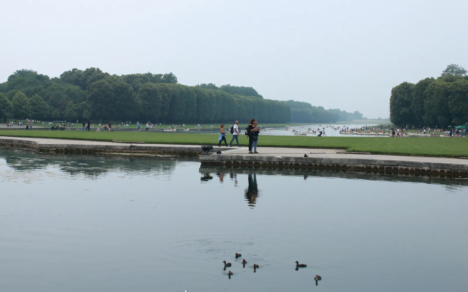 Grand Canal in the Park of Versailles with walkers on the bank and rowing boats in the distance, hazy summer day