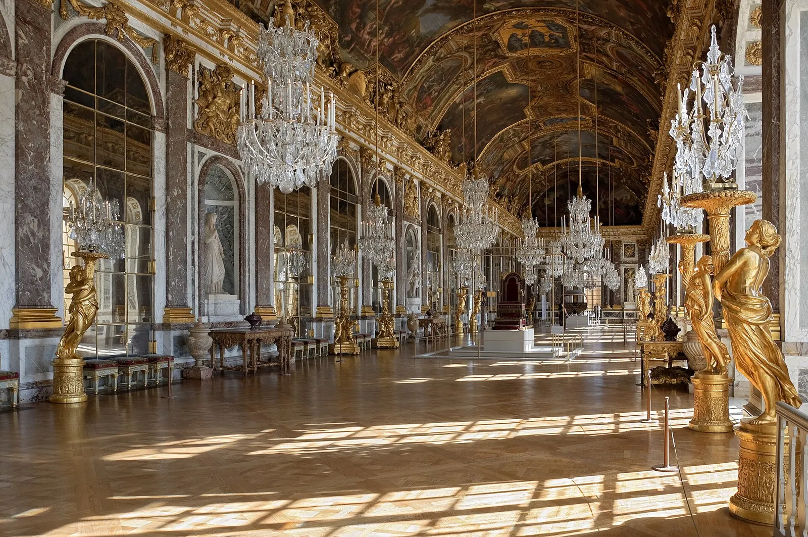 Hall of Mirrors in the Palace of Versailles: a long gallery with arched mirrors on one side, crystal chandeliers, gilded figures and a painted vaulted ceiling, lit by sunlight on a parquet floor