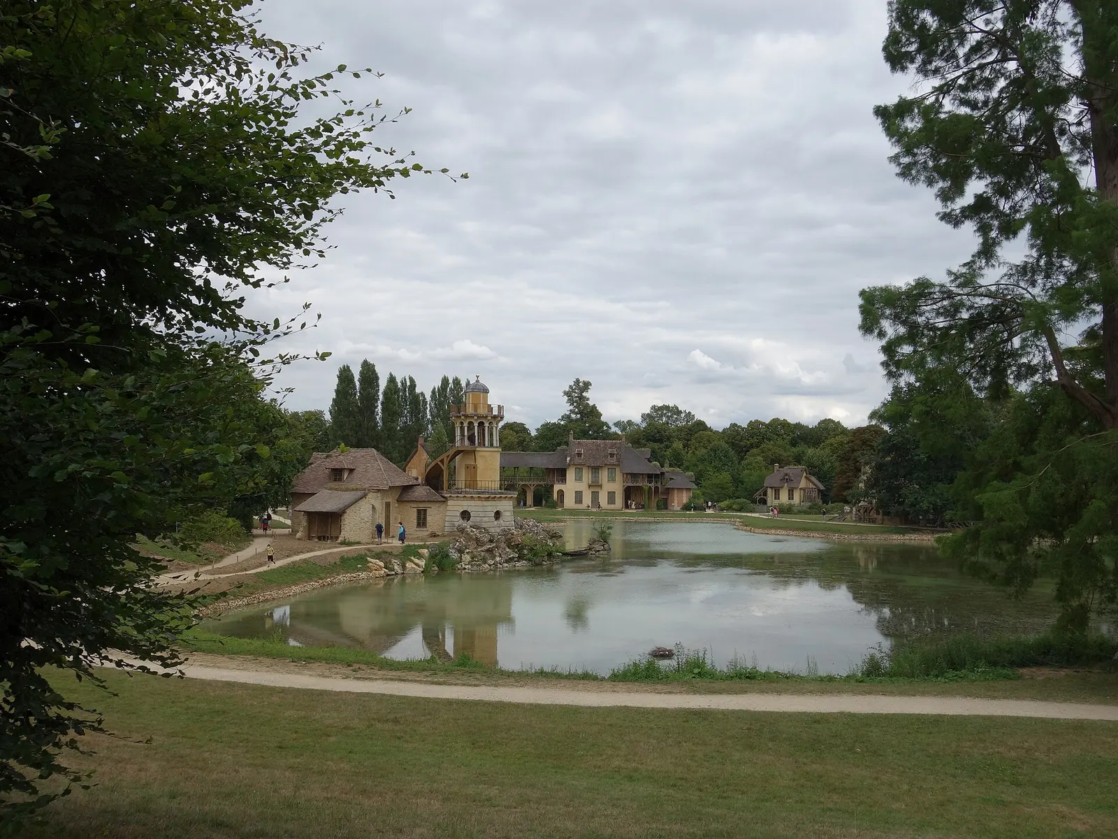 Queen's Hamlet in the Estate of Trianon: farm buildings and a small tower beside a still pond, seen from the lawn under a cloudy sky