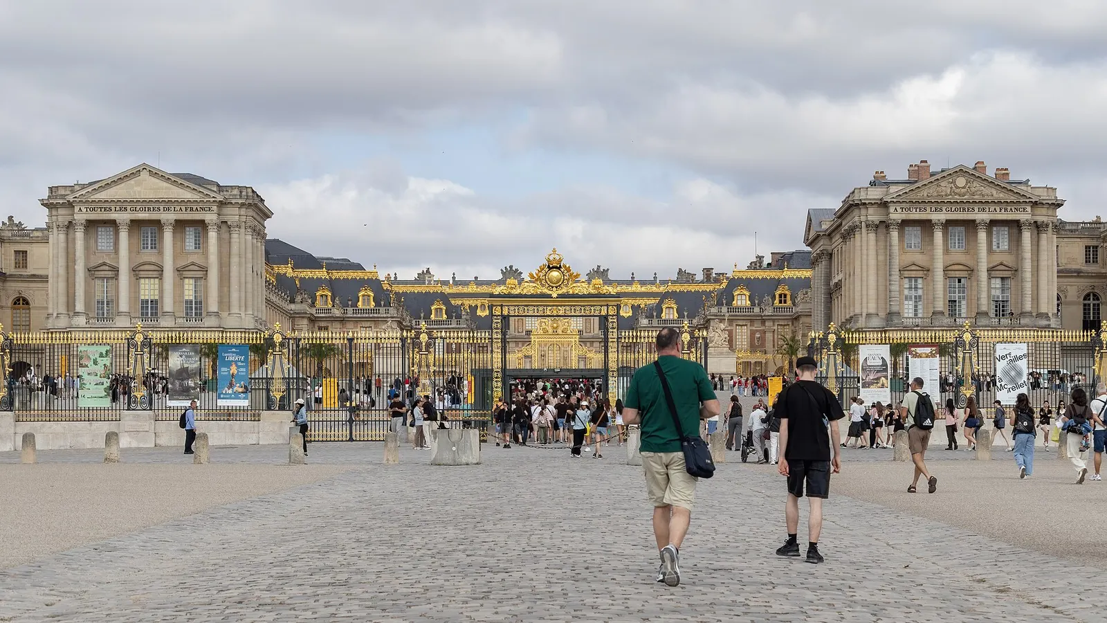 Gilded gate and the Cour d'Honneur of the Palace of Versailles with visitors walking toward the entrance, cloudy day