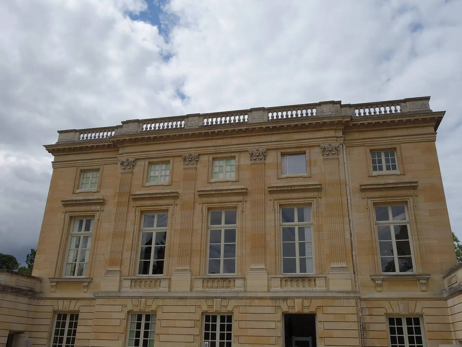 Stone facade of the Petit Trianon with tall windows and a balustrade under a cloudy sky, Estate of Trianon